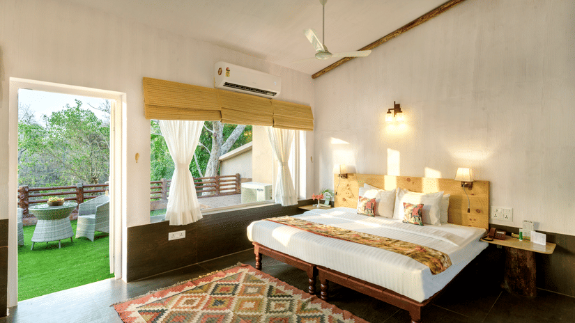 Premier Riverview Suite Twin at The Riverwood Forest Retreat, Pench featuring double size bed, bedside table with telephone, a window with blinds and a curtain, and a balcony with seating area.