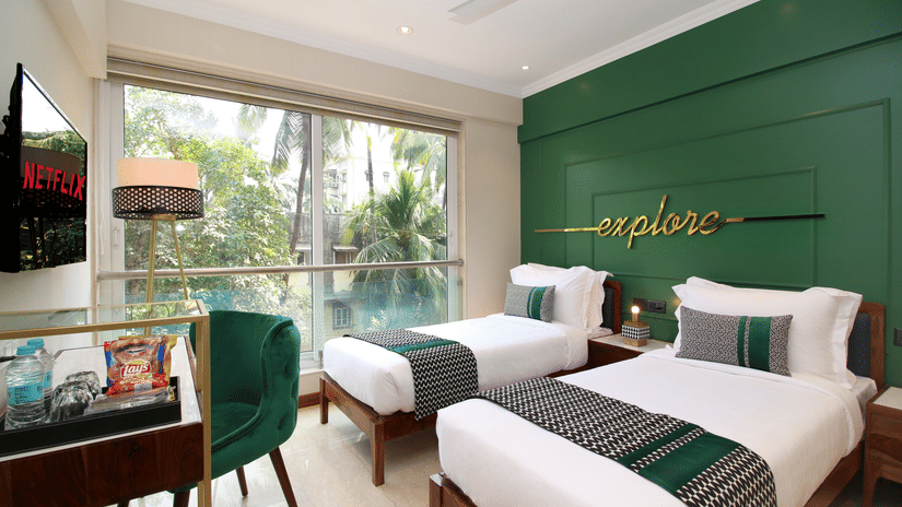 Twin beds facing the TV on the wall with a table and chair below, and large windows by the bed at Theory9 - Premium Srervice Apartments, Bandra