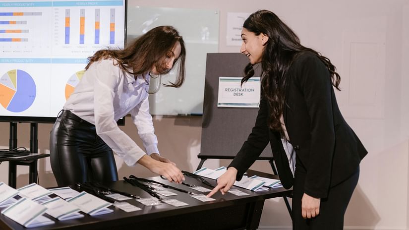 Two women discussing materials at a desk with charts displayed in the background.