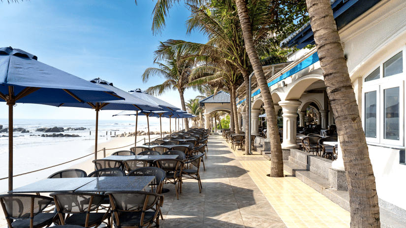 Outdoor beachfront restaurant with shaded seating, palm trees, and ocean views along the shoreline