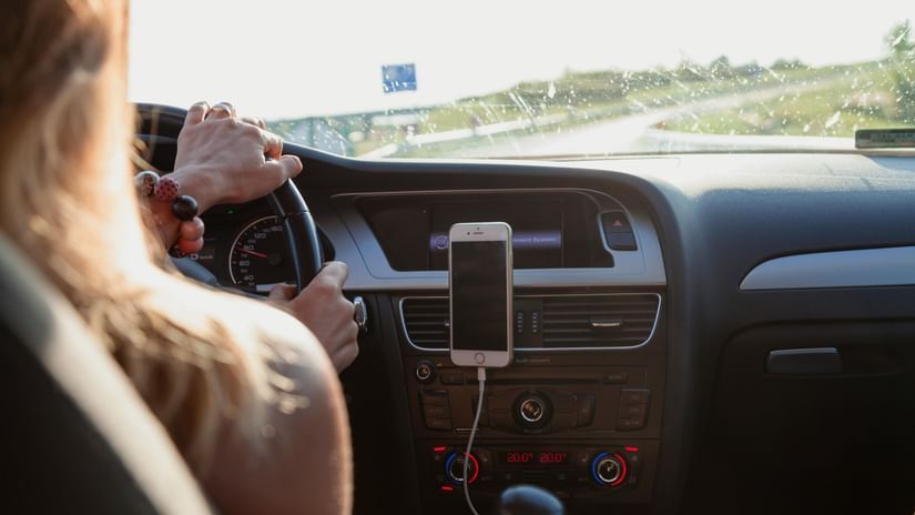 Person drives a car with a mobile phone mounted on the dashboard, view of the steering wheel, dashboard, and road ahead, related to V FIVE HOTEL.
