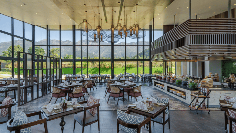 The Brahmkamal Restaurant at Vandhara Sarovar Premiere, Patkote, featuring dark wood tables, floor-to-ceiling glass walls, and hanging lantern lights.
