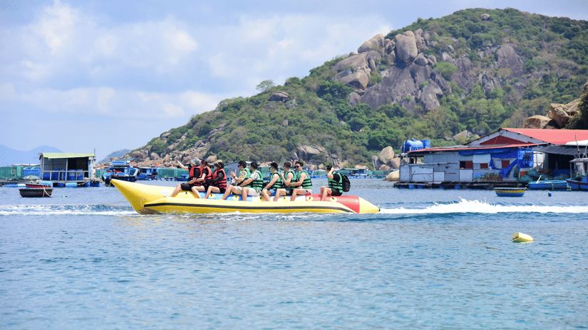 Tourists wearing life jackets enjoy a banana boat ride across coastal waters near rocky hills.