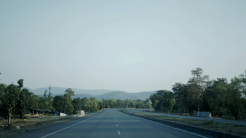 A wide, open highway stretching through a quiet rural landscape with trees on either side and distant hills under a clear sky.