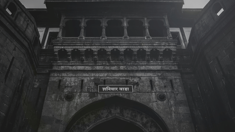 A stone gateway with an arched entrance, balconies with railings above, and a signboard mounted over the arch against the sky.