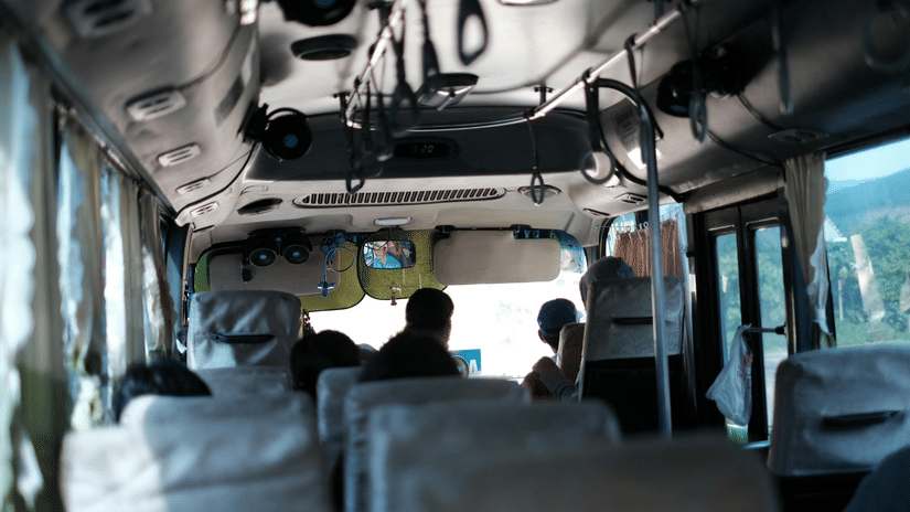View from the back of a bus with passengers seated, showing grey headrests and overhead handrails in soft light.