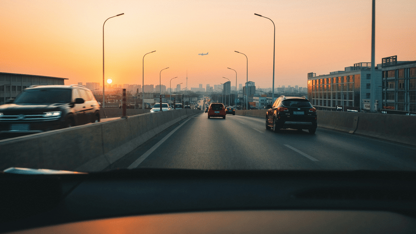 A view from inside a car showing a road with vehicles, streetlights, buildings on both sides, and the sky during sunset.
