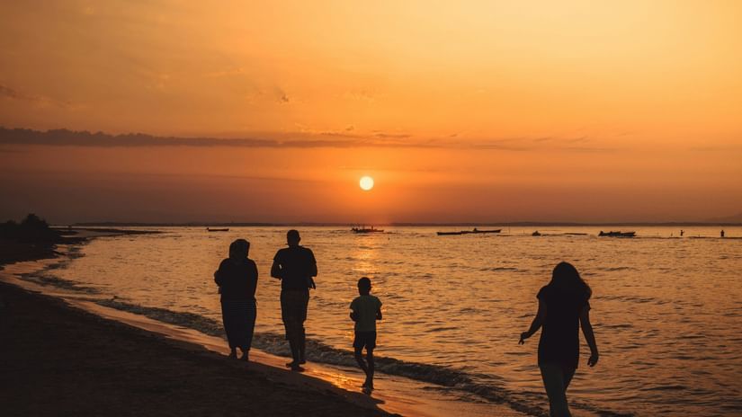  A silhouette of four people, walking along a sandy beach at sunset, with the sun low on the horizon casting an orange glow over the calm water and a few distant boats.