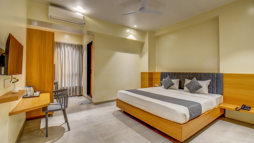 A well-lit Deluxe Room featuring a plush bed facing a wall-mounted TV, a work desk near the wardrobe, and an air-conditioner at VITS Select Nashik Hotels & Resorts.