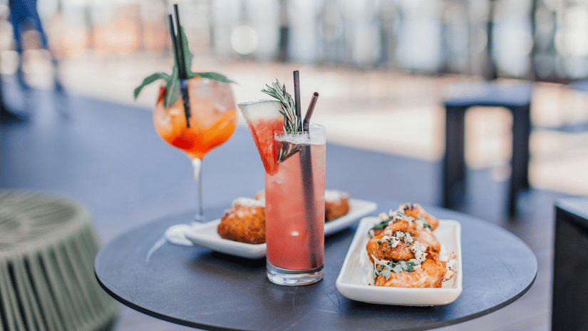 A close up of a small, round, dark outdoor table holding 2 colourful cocktails with garnishes, next to 2 small white plates with snacks