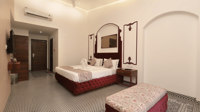 Elegant hotel bedroom with double bed, patterned bedding, bedside lamps, decorative mirror with arches, cushioned bench, tiled floor, and warm ambient lighting.