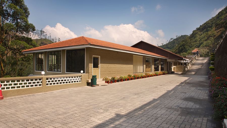 A low-angle shot of a tan building with a red tile roof, a paved walkway, and a mountain backdrop - Abad Copper Castle, Munnar