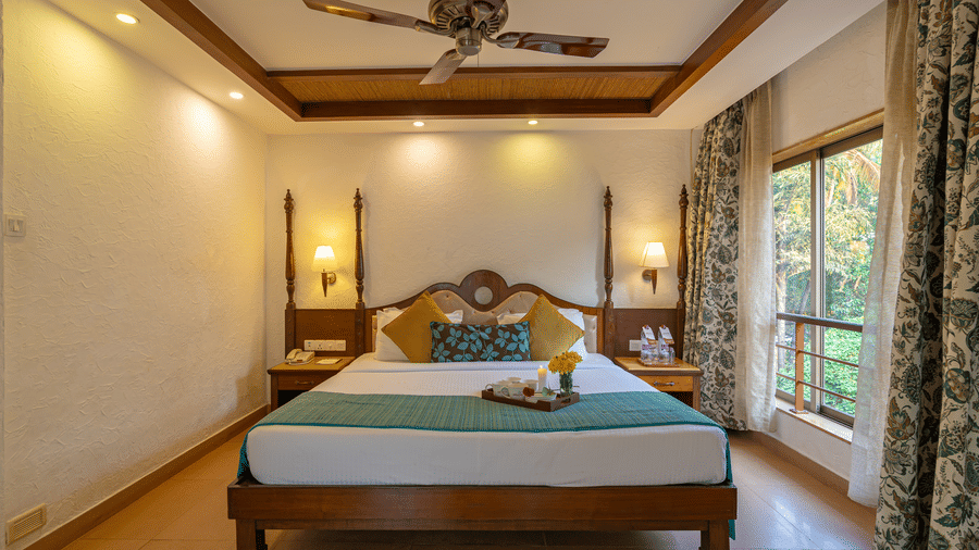An image of a hotel room featuring a beautifully decorated bed with a tray of cups, plates, and flowers, complemented by a large window, a fan, and table lamps.