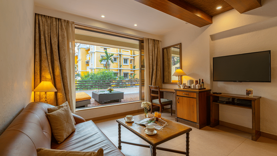 An image of a living room with a brown sofa, small coffee table, TV, storage cabinet, tea and coffee maker, and a balcony with a seating area.