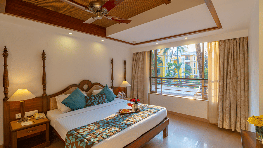 An image of a room featuring a neatly made bed, a fan, two table lamps, side drawers, and a large window.