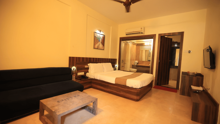 A hotel room at the Adamo The Resortfeaturing a bed, a sofa, centre table, console, table, wall mounted TV, AC, and a glass window with a view of the bathroom.