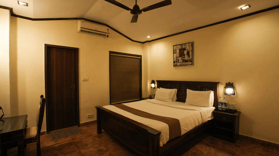 Interior view of a bedroom at Adamo The Village with a double bed,  side tables with lamps, a work desk, and a dark ceiling fan.