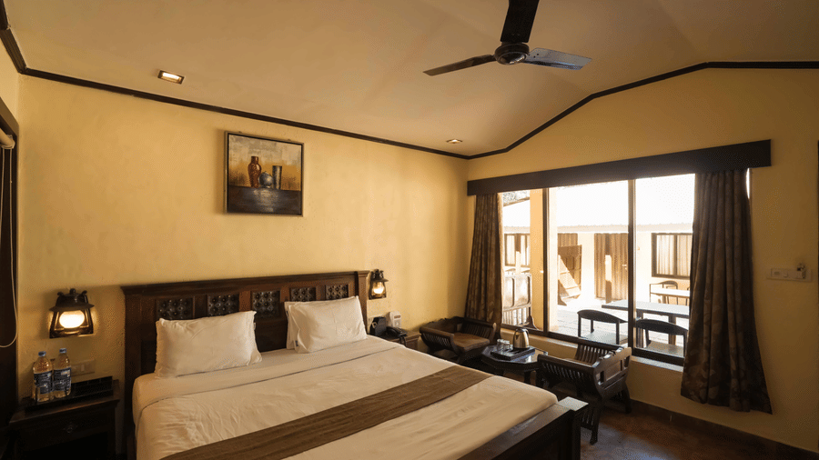 A room at Adamo The Village with a double bed, a large window, a seating area, and a side table with a lamp.