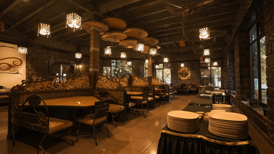 The dining area in Adamo The Village with neatly arranged tables and chairs, wooden ceiling with multiple overhead lamps, and primitive-style decor.