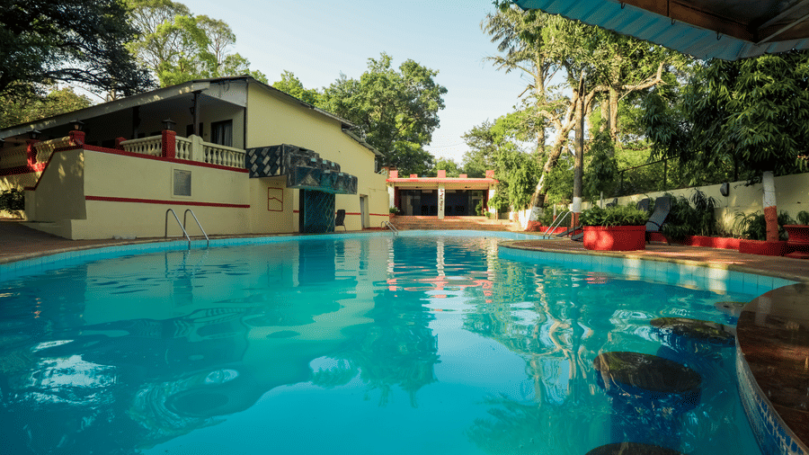 A clear, bright blue swimming pool winding alongside resort buildings and shaded by lush green trees.