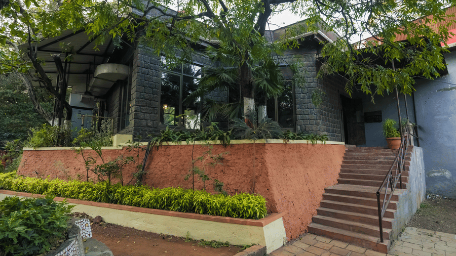 A close view of a dark-painted cottage at Adamo The Village with a small staircase, nestled amidst lush greenery and trees.