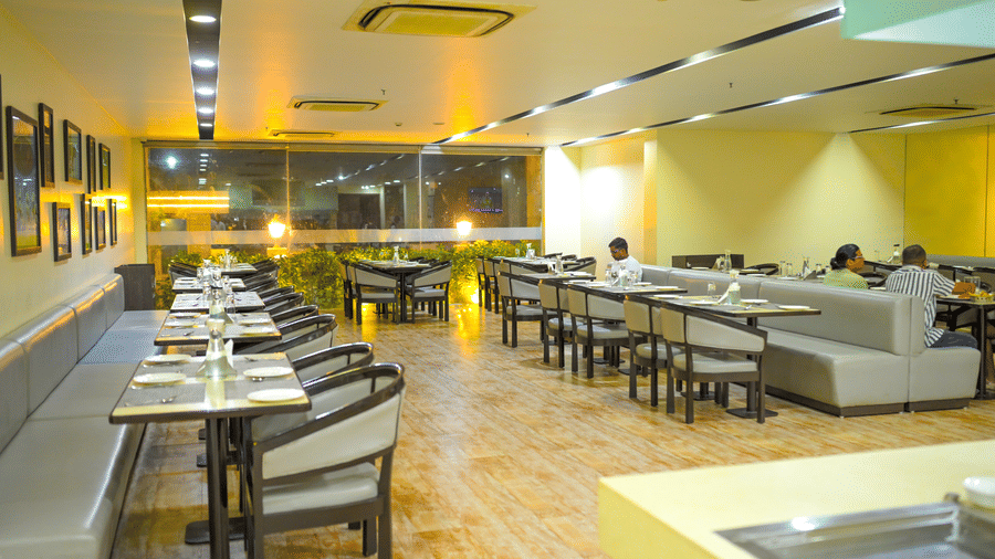 A restaurant dining room at Ambar Sarovar Portico, Gandhidham features neatly arranged tables, chairs, and cushioned booth seating along the wall.