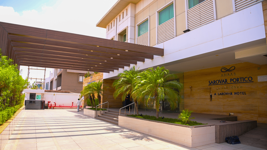 A daylight view of the main building exterior at Ambar Sarovar Portico, Gandhidham showing a covered canopy entrance, steps, and small palm trees.
