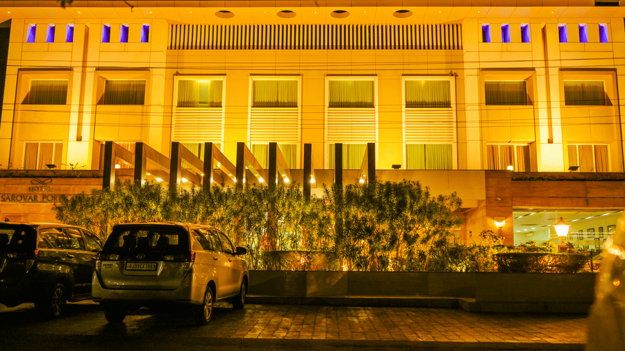 The exterior facade of Ambar Sarovar Portico, Gandhidham is illuminated at night with yellow lights above parked cars on the driveway.