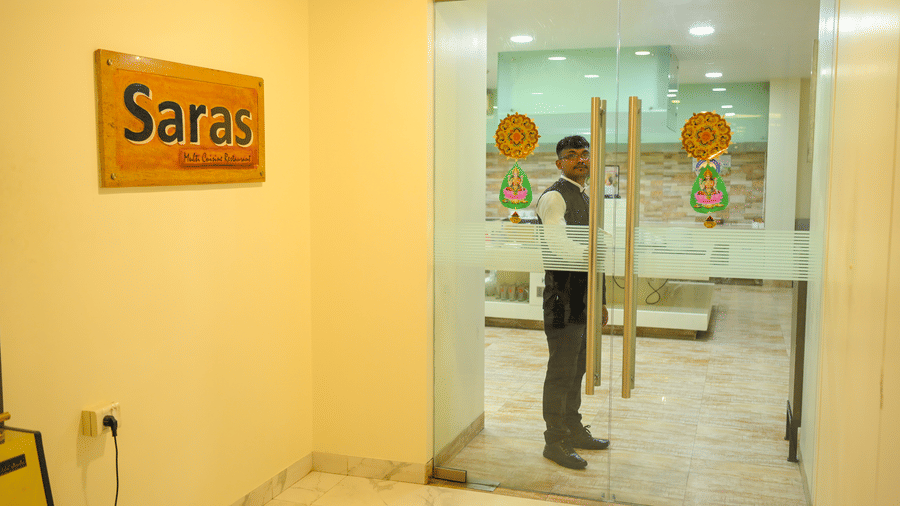 An entrance to Saras restaurant at Ambar Sarovar Portico, Gandhidham features a glass door, a staff member, and a wooden signboard on the wall.
