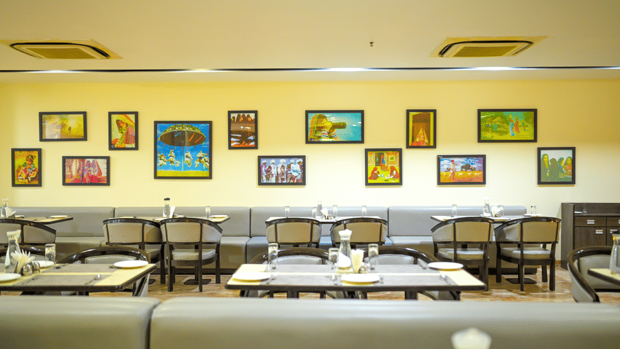 A dining space at Ambar Sarovar Portico, Gandhidham shows a yellow wall decorated with multiple framed paintings, dining tables, and chairs.