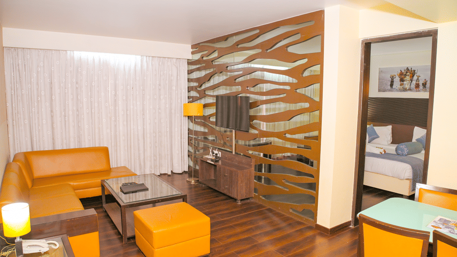A suite living room at Ambar Sarovar Portico, Gandhidham contains an L shaped sofa, a coffee table, and a television mounted on a patterned wooden wall.