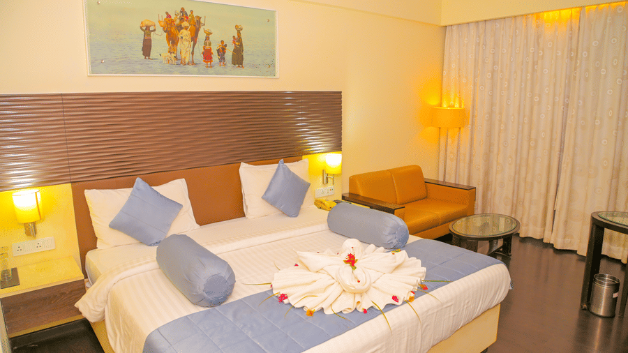 A hotel bedroom at Ambar Sarovar Portico, Gandhidham shows a large king size bed decorated with folded towels, a sofa, and a wall painting.