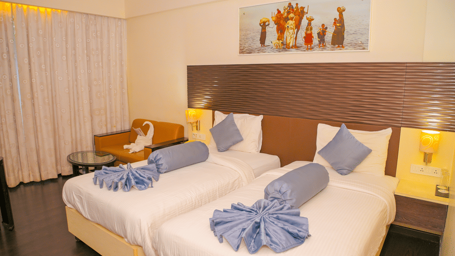 A hotel room at Ambar Sarovar Portico, Gandhidham features 2 separate single beds, pillows, a wall painting, a small armchair, and curtains.