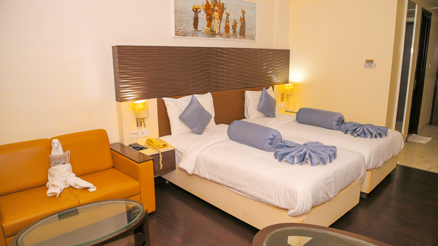 A suite room at Ambar Sarovar Portico, Gandhidham is arranged with 2 separate beds, pillows, a couch, glass coffee tables, and a dark wooden floor.