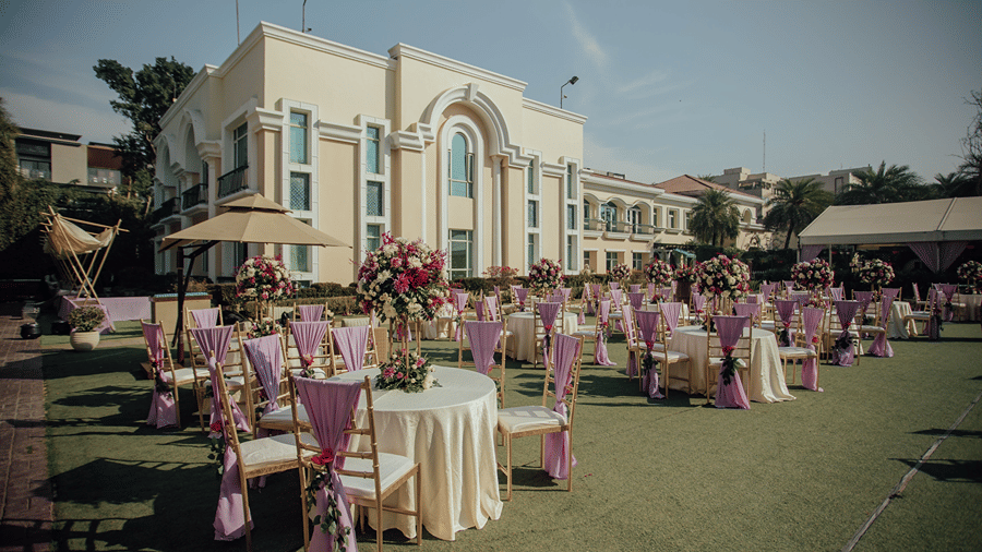 Elegantly decorated outdoor garden lawn setup with round banquet tables and draped chairs in front of a hotel building.