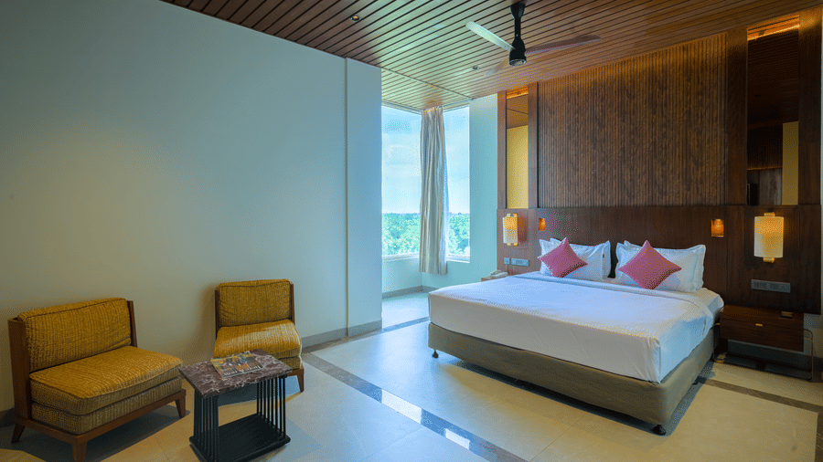 Hotel bedroom featuring a king size bed, two lounge chairs, wooden ceiling, warm lighting and a dark wood feature wall.