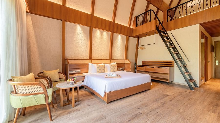 An image of attic style bedroom with a king size bed, a wooden seating area, a flat TV screen, a table counter top, and a  step ladder to reach the attic