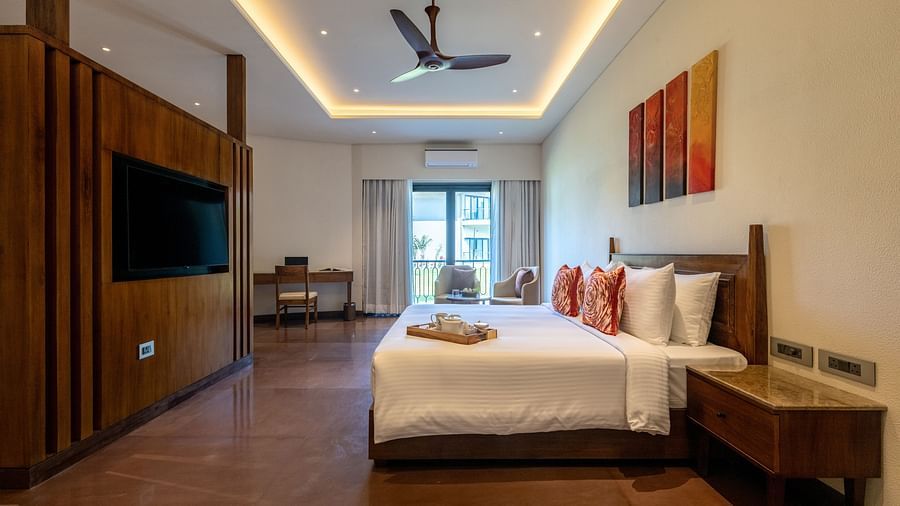 An image of a bedroom with a king size bed where tea tray is placed, a flat TV screen on top of a table counter top, a seating area at one corner, and a  large window at one side