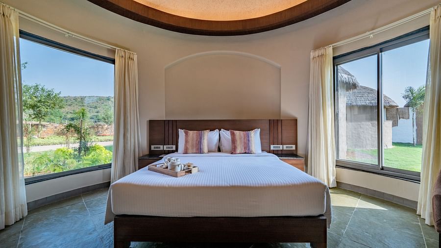 An image of a bedroom with king size bed, with bedside table on either side along with two large windows with curtains removed to let the natural light in