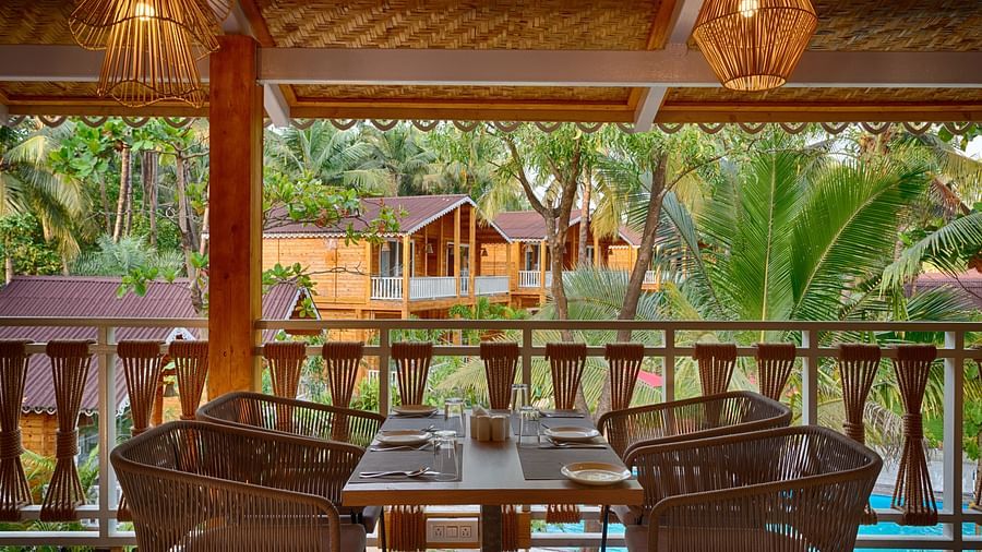 View from a covered dining area with woven chairs, a set dining table, overlooking wooden cabins among palm trees Aralea Beach Resort by Stone Wood, Morjim