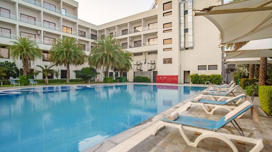 Wide shot of the swimming pool deck, showing sun loungers and the multi-story white hotel building at Arawali Ananta Elite, Jaipur.