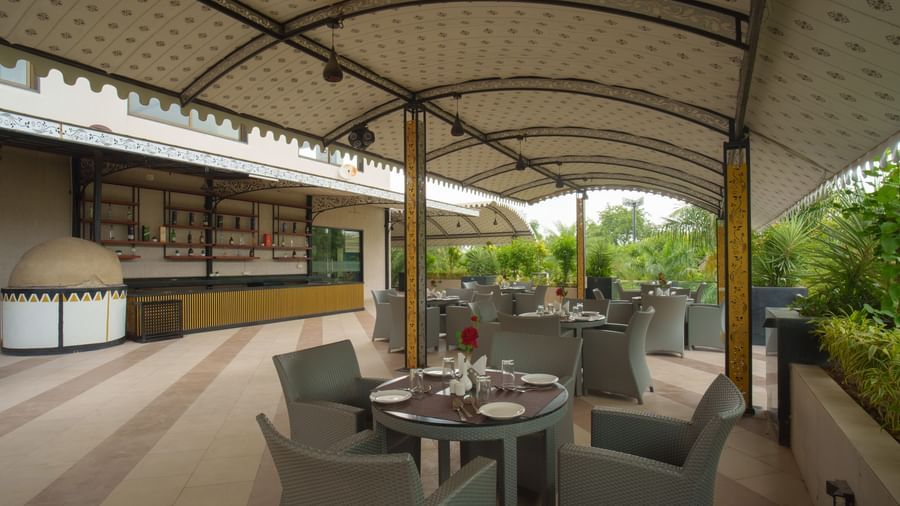 View of the covered outdoor restaurant space showing dining tables, chairs, and a service counter in the background at Arawali Ananta Elite, Jaipur.