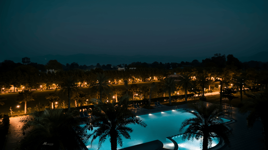 An outdoor swimming pool at Arawali Ananta Elite Jaipur is lit up at night with palm trees surrounding it.