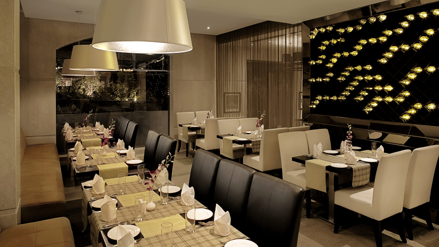 Modern dining room at Bahula restaurant in Barsana Boutique Hotel, Kolkata, featuring long set tables with elegant chairs and soft ambient lighting from large overhead lamps.