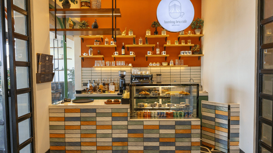 Cafe front at Hotel Bawa, Udaipur displaying a tiled counter, glass food cabinet, espresso machine, wall shelves, and circular overhead logo sign.