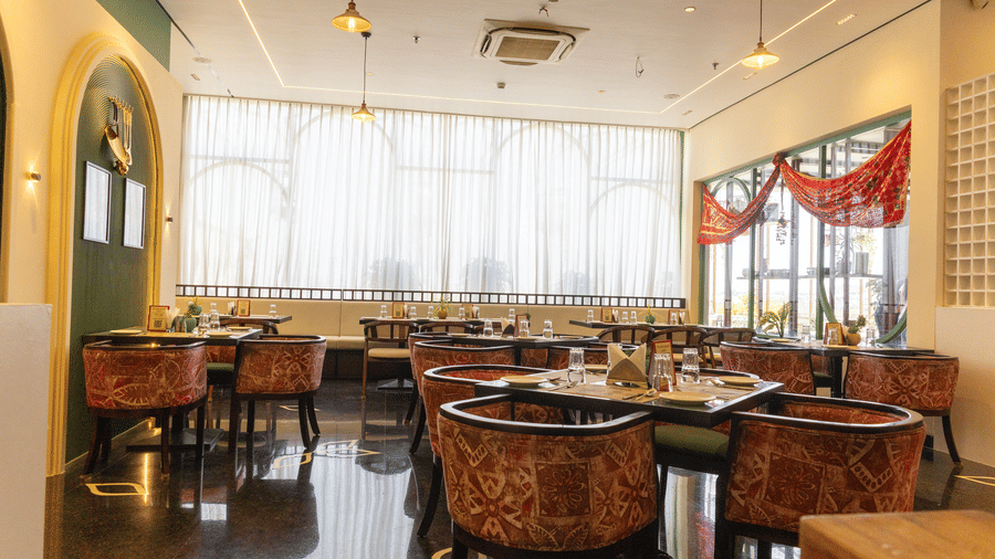 A restaurant at Hotel Bawa, Udaipur showing dining tables, patterned armchairs, sheer curtained windows, hanging lights, and polished flooring.