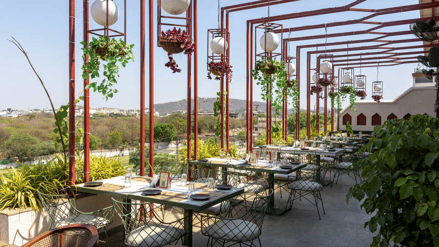 Outdoor rooftop dining section at Hotel Bawa, Udaipur showing metal canopy structures, hanging plant baskets, dining tables, and scenic views.