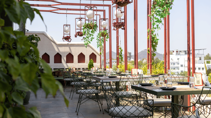 Outdoor rooftop terrace at Hotel Bawa, Udaipur featuring dining tables, chairs, metal overhead framework, hanging lantern lamps, and greenery.