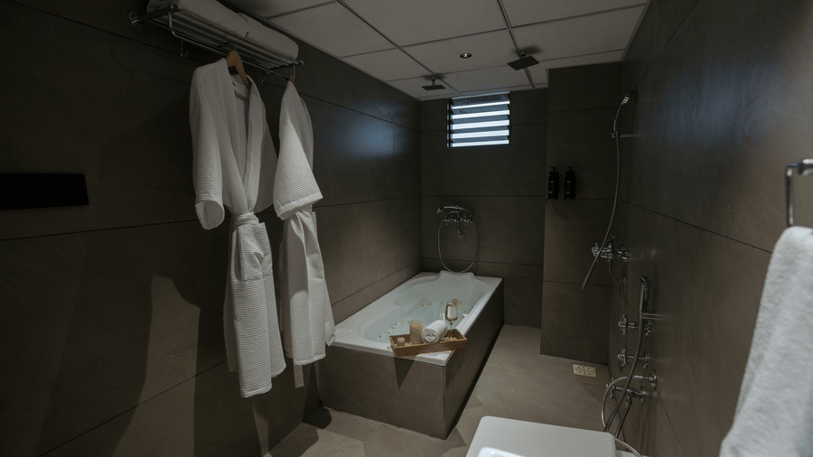 Modern hotel bathroom at Biznotel by Pride Surat with a deep soaking bathtub, plush white bathrobes, and sleek dark walls.