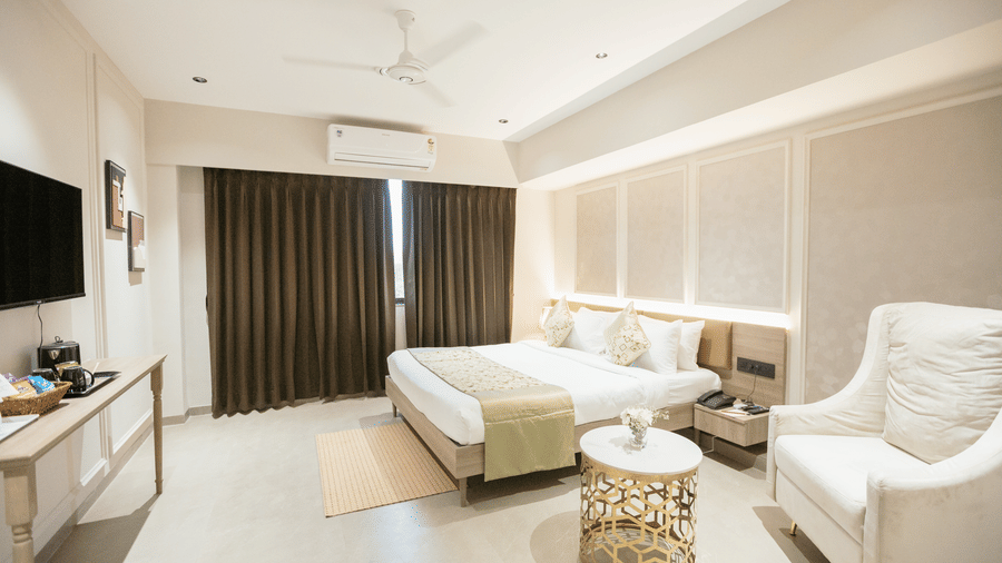 Bright and spacious guest room at Biznotel by Pride Surat featuring a large bed, seating armchair, TV setup, and floor-to-ceiling window curtains.